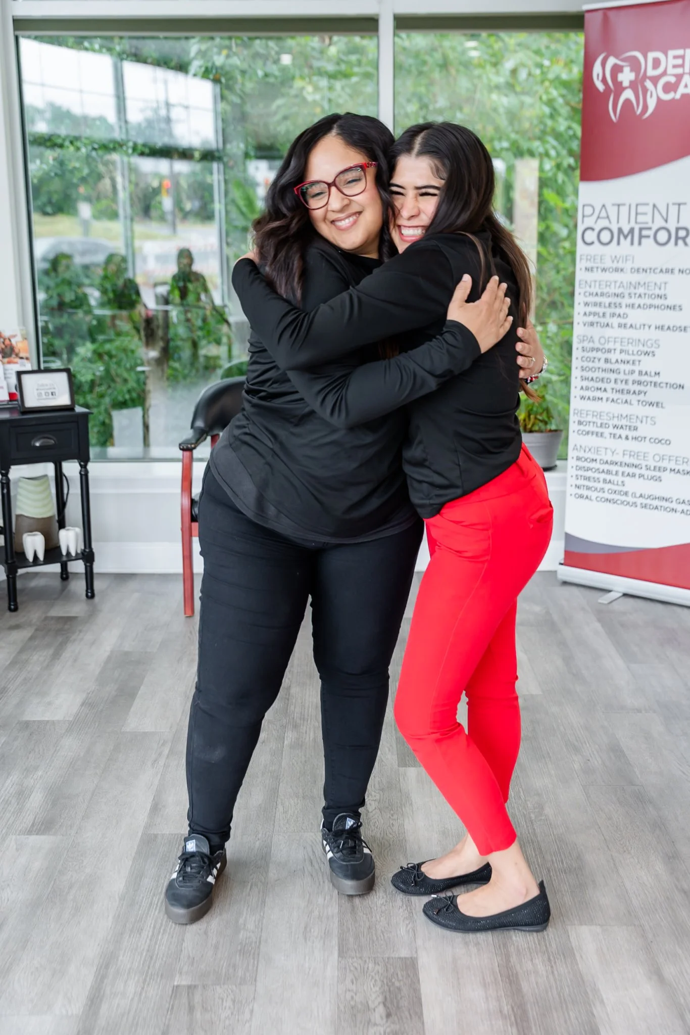 Two team members hugging in a modern dental office waiting area with a Dentcare Now branded banner visible
